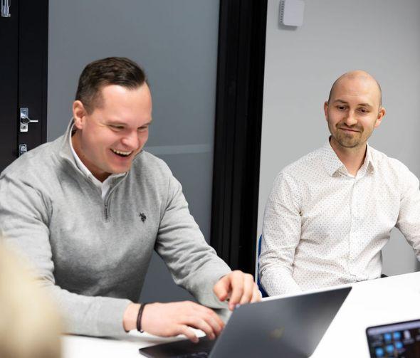 Harmaapaitainen mies näpyttelee tietokonetta ja valkopaitainen mies seuraa vieressä