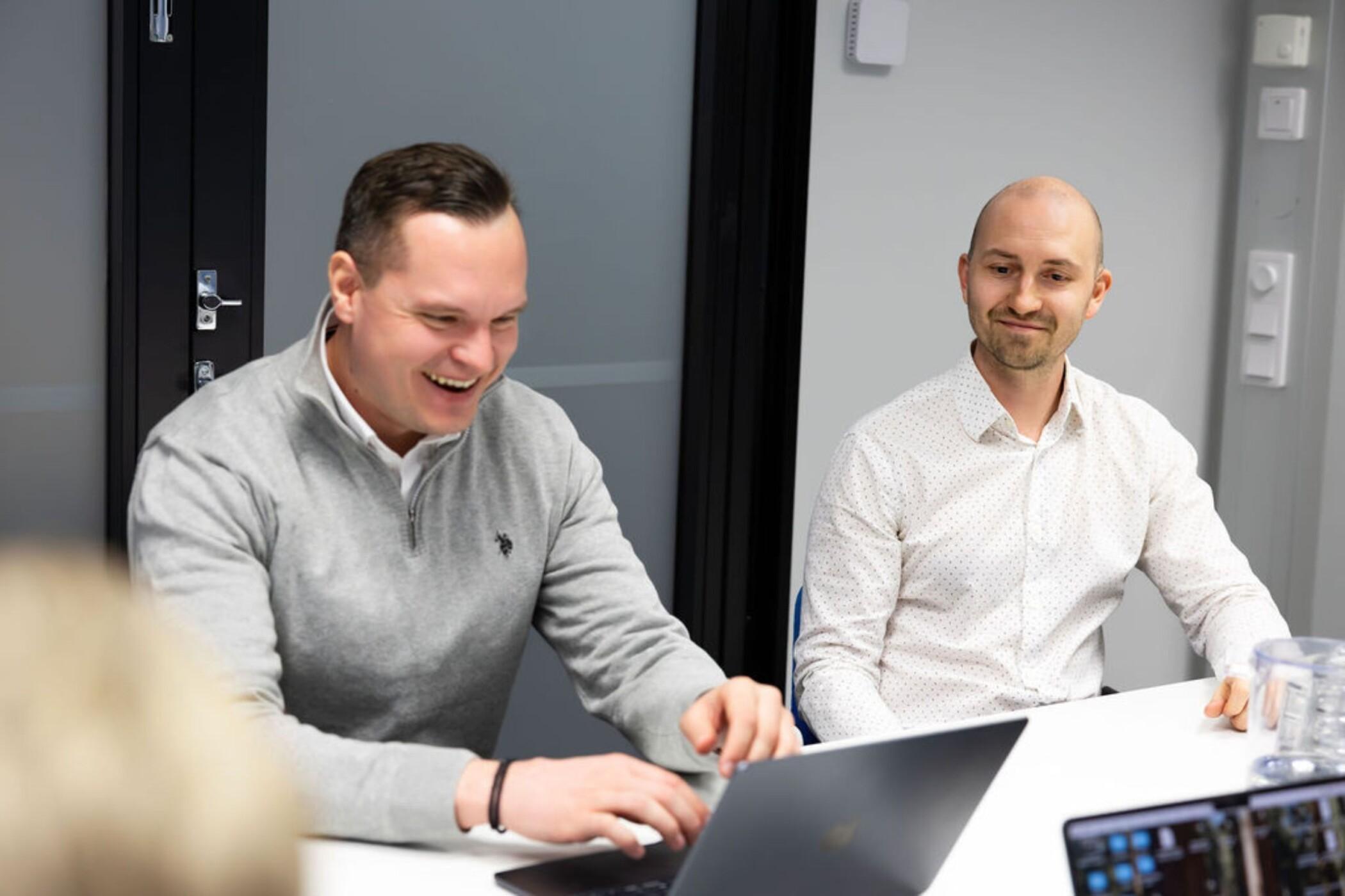 kaksi miestä neuvottelutilassa ja toinen näppäilee tietokonetta